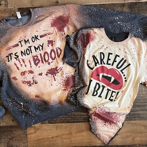 May include: Two Halloween-themed shirts. A dark grey sweatshirt with the text "I'M OK IT'S NOT MY BLOOD" and blood splatter design. A short-sleeved shirt with "CAREFUL... I BITE!" and vampire lips.