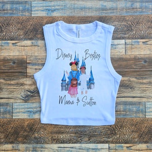 May include: White sleeveless crop top featuring a graphic print of two people standing in front of a blue castle. The text "Disney Besties Mama & Sutton" is printed on the shirt.