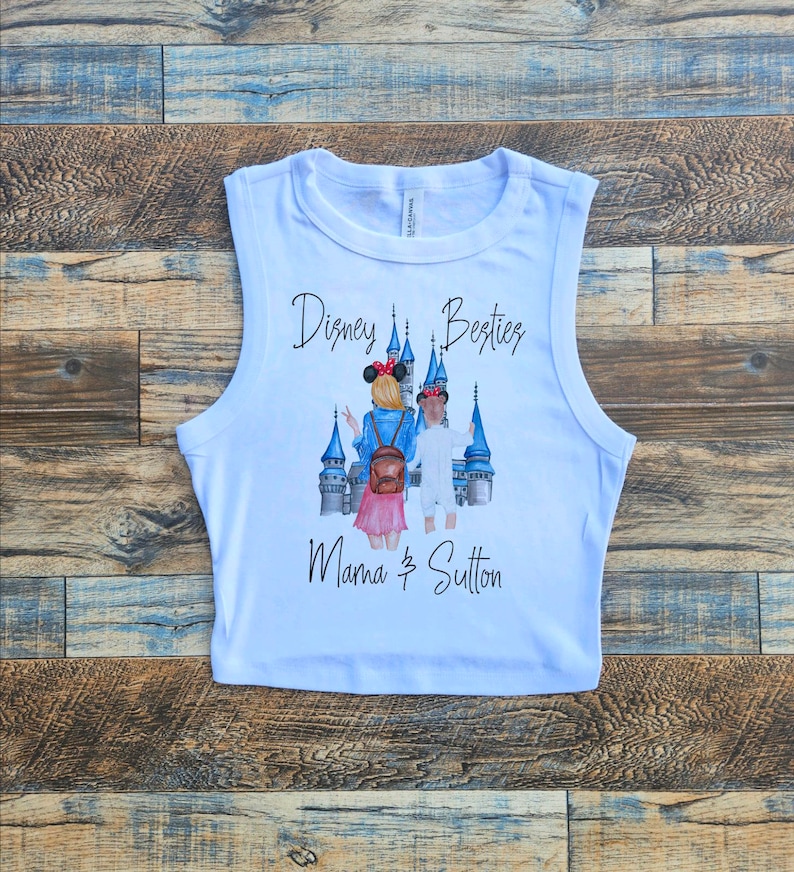 May include: White sleeveless crop top featuring a graphic print of two people standing in front of a blue castle. The text "Disney Besties Mama & Sutton" is printed on the shirt.