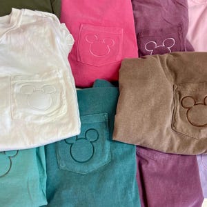 May include: A selection of colourful t-shirts with a pocket featuring a Mickey Mouse design. The shirts are in various colours, including white, pink, teal, brown, and purple. The pocket design is embroidered in a matching or contrasting colour.