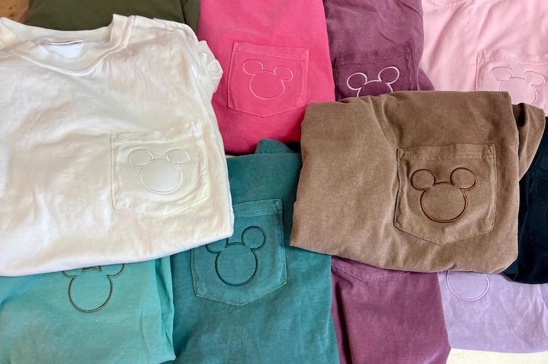 May include: A selection of colourful t-shirts with a pocket featuring a Mickey Mouse design. The shirts are in various colours, including white, pink, teal, brown, and purple. The pocket design is embroidered in a matching or contrasting colour.