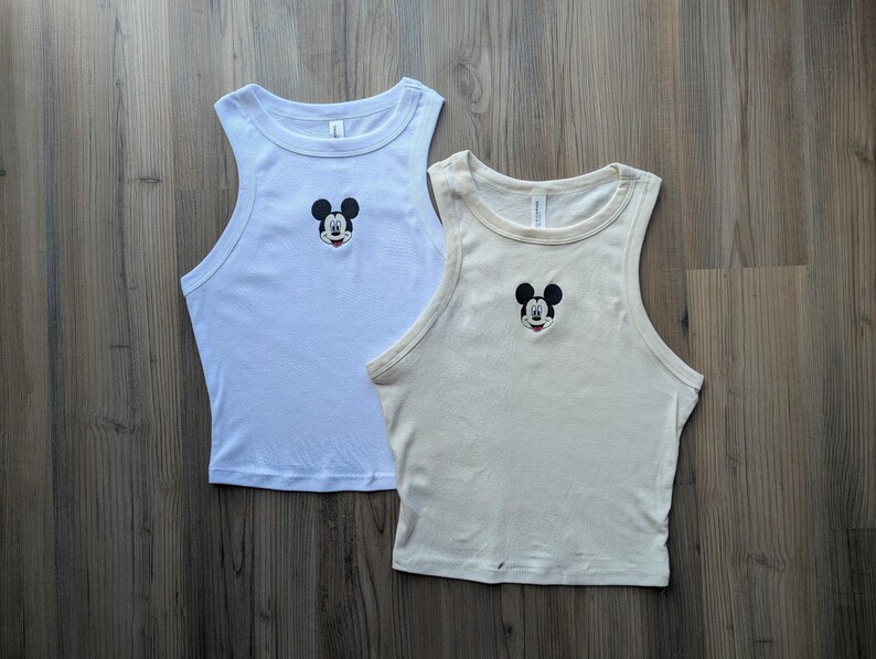 May include: Two sleeveless tank tops, one light blue and one cream-colored, are displayed on a wooden surface. Each top features an embroidered Mickey Mouse design on the chest. The tops have a classic, casual style.
