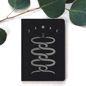 May include: A black notebook with a silver snake design and the word "D.O.D.C." above the snake. The snake is coiled around a circle with a dot in the center.
