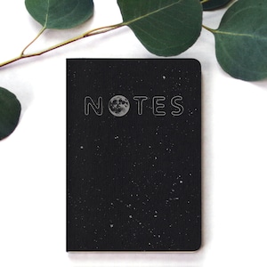 May include: A black notebook with a white outline of the word "NOTES" and a silver moon graphic. The notebook has a speckled, starry night design.