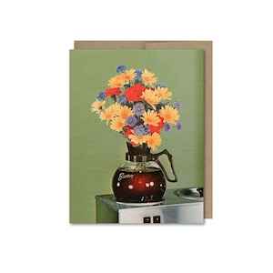 May include: A glass coffee pot with a bouquet of yellow, orange, and blue flowers inside. The pot is sitting on a silver coffee maker.