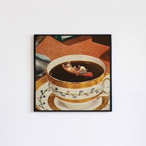 May include: A white teacup with gold trim and a floral pattern sits on a saucer with a gold rim and blue floral accents. The teacup is filled with dark brown liquid and a red canoe with two people paddling is floating on top.