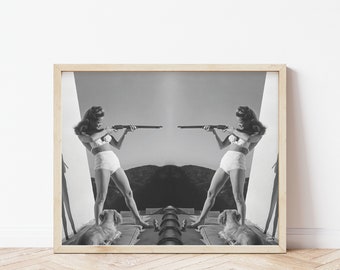 May include: A black and white photo of a woman in a swimsuit holding a rifle in each hand. The woman is standing in front of a mirror, creating a symmetrical image. Two dogs are lying on the ground in front of her.