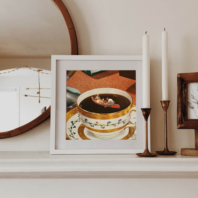 May include: A white framed print of a surrealist image of a teacup filled with dark liquid with a small red canoe with two people in it floating on top.