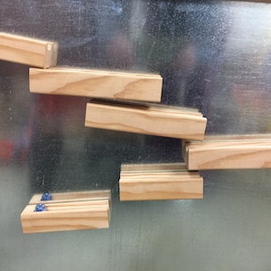 May include: Wooden blocks arranged in a cascading pattern, attached to a metallic surface. The blocks are rectangular, with a natural wood finish. Two small blue marbles are visible on one of the blocks. The arrangement suggests a playful or decorative purpose.