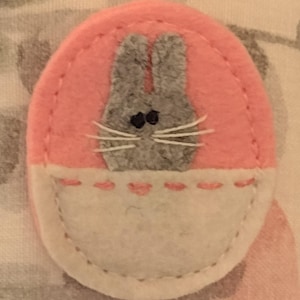 May include: A felt Easter egg-shaped applique with a gray bunny face. The bunny has black eyes and white whiskers. The egg is pink with a white pocket at the bottom, stitched with pink thread.