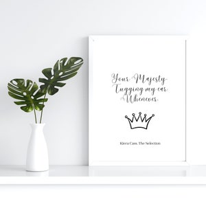 May include: A white framed print featuring the text "Your Majesty Tugging my ear Whenever" and a crown illustration. The print is by Kiera Cass, The Selection. A white vase with green leaves is to the left.