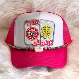 May include: A pink trucker hat with a white front panel featuring the text "TILE JUNKIE" and images of Mahjong tiles. The hat has a curved brim and a beaded accent.
