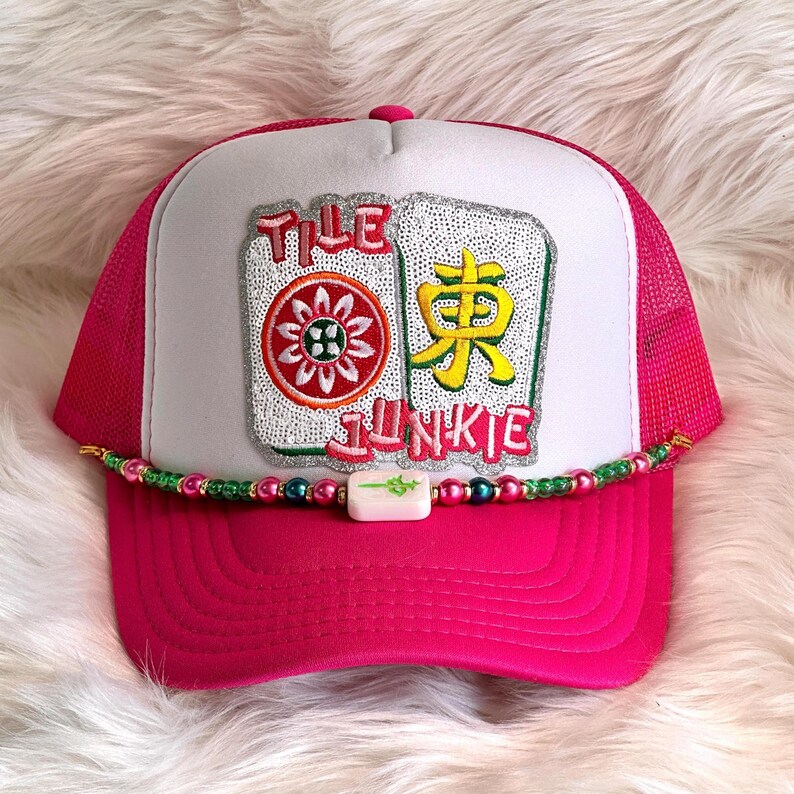 May include: A pink trucker hat with a white front panel featuring the text "TILE JUNKIE" and images of Mahjong tiles. The hat has a curved brim and a beaded accent.