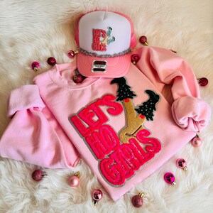May include: A pink sweatshirt and trucker hat with the text "Let's Ho Girls." The sweatshirt has a sequined design of a figure with Christmas trees for legs. The hat is pink and white with a silver brim and the same text.