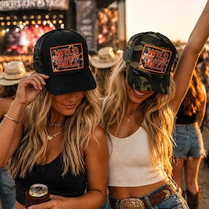 May include: Two women wearing trucker hats with the text "Wild as Hell" on a square patch. One hat is black, the other is camouflage. The hats have a mesh back and a curved brim. The women are smiling and appear to be at an outdoor event.