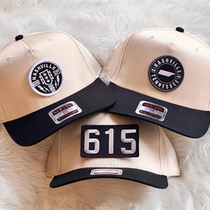May include: Three beige and black baseball caps with patches. Two caps have a Nashville patch with a guitar and lightning bolts. The third cap has a patch with the number "615".
