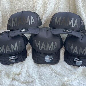 May include: Five black trucker hats with a silver 'MAMA' graphic on the front. The hats are all black with a mesh back and a black adjustable strap.