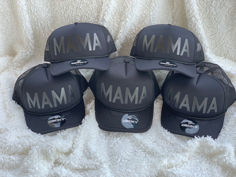 May include: Five black trucker hats with a silver 'MAMA' graphic on the front. The hats are all black with a mesh back and a black adjustable strap.