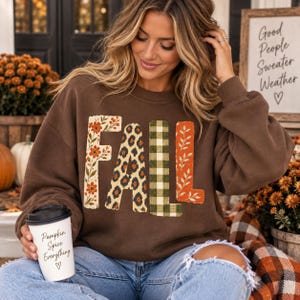 May include: A dark brown crewneck sweatshirt featuring the word FALL in large block letters. Each letter has a unique pattern: the F is floral, the A is leopard print, the L is green and white plaid, and the L is orange with white leaves.