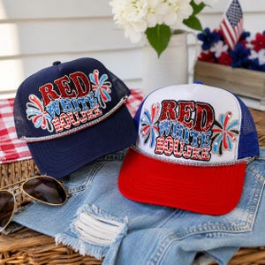 May include: Two trucker hats featuring the text RED WHITE BOUJEE in glittery red and blue lettering with firework graphics. One hat is navy blue with a matching mesh back, while the other has a white front, red brim, and blue mesh back.
