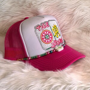May include: A pink trucker hat with a white front panel featuring a colorful embroidered design and the text "TILE JUNKIE". The hat has a pink mesh back and a beaded accent along the brim.
