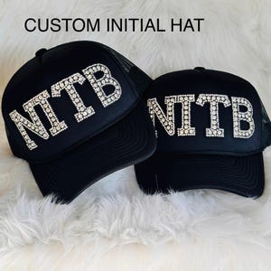 May include: Two black trucker hats with the initials "NTB" spelled out in sparkling rhinestones. The hats have a mesh back and a curved brim. The text "CUSTOM INITIAL HAT" is at the top of the image.