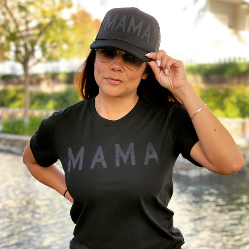 May include: A black t-shirt with the word "MAMA" printed in large, bold letters. The model is wearing a black mesh trucker hat with the same word printed on it.
