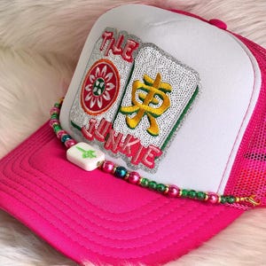 May include: A pink trucker hat with a white front panel and mesh back. The hat features an embroidered patch with the text "THE JUNKIE" and two decorative tiles. A beaded necklace with a white pendant is draped across the hat.