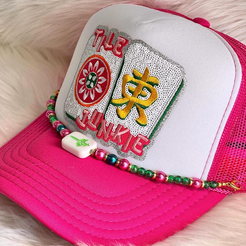 May include: A pink trucker hat with a white front panel and mesh back. The hat features an embroidered patch with the text "THE JUNKIE" and two decorative tiles. A beaded necklace with a white pendant is draped across the hat.