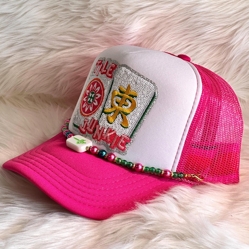 May include: A pink trucker hat with a white front panel. The hat features a colorful embroidered design with the words "TILE JUNKIE" and a beaded accent. The mesh back and brim are bright pink.