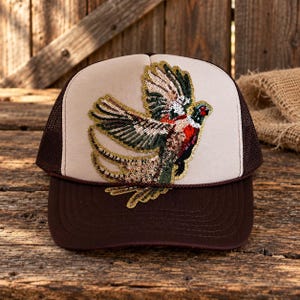 May include: A beige and brown trucker hat featuring a sequined pheasant bird patch on the front panel. The cap has a solid beige front, a dark brown brim, and brown mesh side and back panels with a decorative cord across the base of the crown.