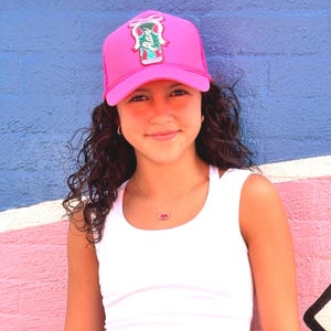 May include: A bright pink trucker hat with a watermelon design and the word "Alani" on the front. The hat is worn with a white tank top and a gold necklace with a pink pendant. The background is a blue and pink painted wall.