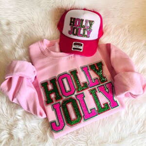 May include: A pink sweatshirt and a red and white trucker hat. The sweatshirt features the words "HOLLY JOLLY" in green and pink glitter. The hat has the same text in a similar style. The items are displayed on a white, fluffy surface.