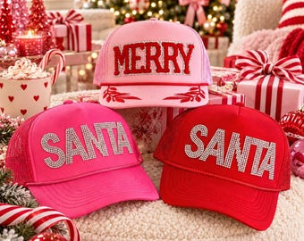 May include: Three baseball caps with mesh backs. The top cap is light pink with red MERRY text and leaf accents. The bottom left cap is bright pink with silver rhinestone SANTA text. The bottom right cap is red with silver rhinestone SANTA text.