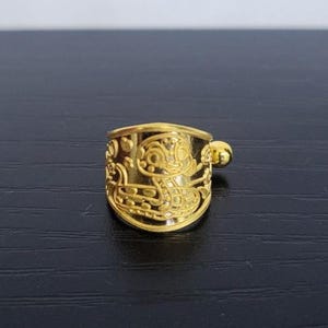 May include: A gold-colored ring with an engraved design featuring a stylized face and other decorative elements. The ring has a rounded shape with a small, spherical detail on the side. The ring is set against a dark background.