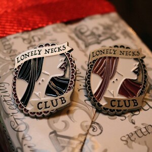 Puede incluir: Dos pines de plata con esmalte negro y rojo. Cada pin presenta una ilustraci&oacute;n estilizada de la cara de una mujer con el texto "Lonely Necks Club".