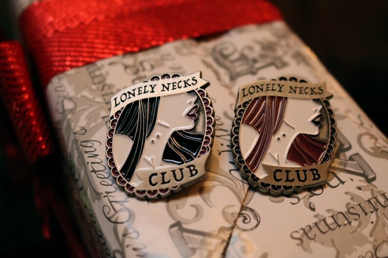 Puede incluir: Dos pines de plata con esmalte negro y rojo. Cada pin presenta una ilustraci&oacute;n estilizada de la cara de una mujer con el texto "Lonely Necks Club".
