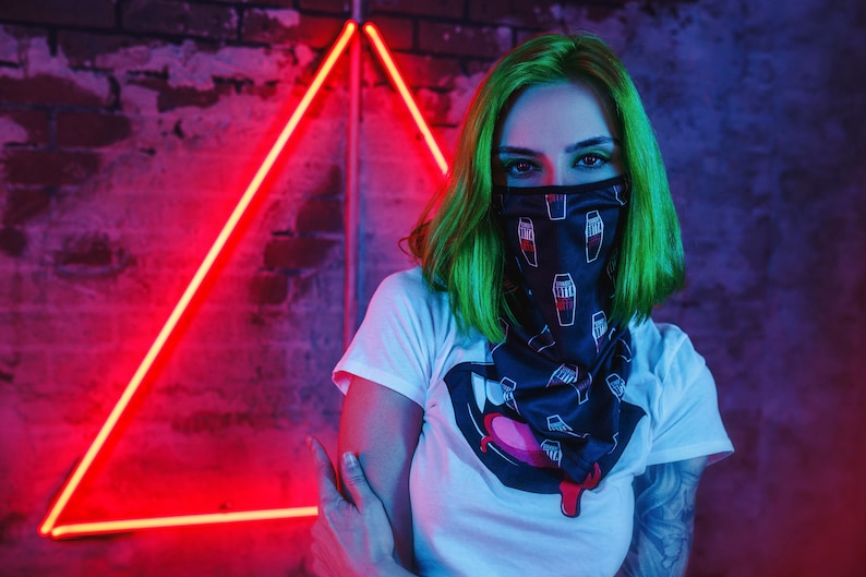 May include: A person wearing a white t-shirt with a black and white graphic print of a mouth with a pink tongue and a black bandana with a white print of coffins. The person has green hair and is standing in front of a red neon triangle.