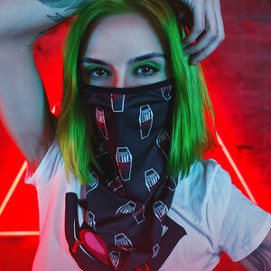 K&ouml;nnte beinhalten: Eine Person mit gr&uuml;nen Haaren tr&auml;gt ein schwarz-wei&szlig;es Bandana mit einem sich wiederholenden Muster aus S&auml;rgen und dem Text "Straight Outta". Das Bandana bedeckt Mund und Nase der Person. Die Person tr&auml;gt ein wei&szlig;es T-Shirt mit einer roten und schwarzen Grafik eines tropfenden Herzens. Die Person hat ein Tattoo eines Pfauenfeders auf ihrem Arm.