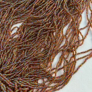 May include: Close-up of a large quantity of small, iridescent seed beads. The beads are primarily brown with hints of gold, purple, and green, creating a shimmering effect. The beads are strung together, suggesting their use in jewelry or craft projects.