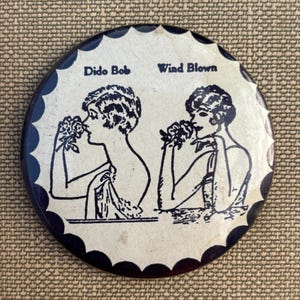 May include: A circular pinback button featuring a cream-colored center with a dark blue scalloped border. The design displays two black line-art illustrations of figures with short hair, each holding a flower. The text reads Dido Bob and Wind Blown.