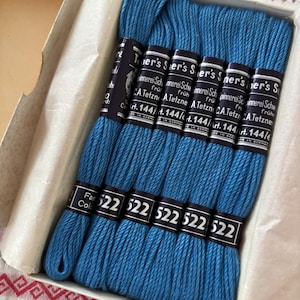 Pode incluir: Uma caixa de fios de bordar azuis. Cada meada é rotulada com o número 522 e o nome da marca. A caixa é branca e tem um forro de papel. O fio é de um tom azul vibrante.