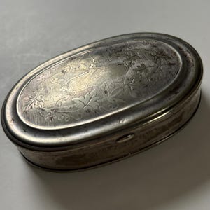 May include: An oval-shaped silver-toned metal box with a hinged lid. The top surface features an intricate engraved pattern of leaves and vines surrounding a central blank cartouche. The metal has a weathered, aged patina finish.