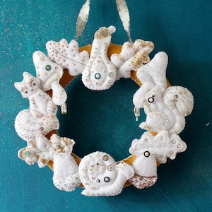 Może przedstawiać: Biały wieniec filcowy ozdobiony haftowanymi leśnymi stworzeniami, w tym wiewiórką, ślimakiem i grzybem. Wieniec jest ozdobiony złotymi i srebrnymi cekinami i koralikami.