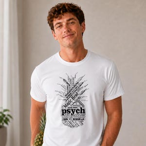May include: A white short-sleeved t-shirt featuring a black graphic print in the shape of a pineapple. The graphic is composed of various words and phrases, including "Magic Head" and "psych" in a prominent, bold, black font style.
