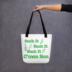 Puede incluir: Bolso tote blanco con asas negras. El bolso tiene la frase "Suck It" en verde, con símbolos de notas musicales y las palabras "C'mon Son". El bolso se sostiene sobre un fondo gris.