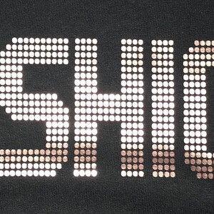 May include: The word FASHION! is displayed in a bold, block-letter font on a solid black background. The letters are constructed from a grid of small, metallic, copper-colored square studs that create a reflective, textured appearance.