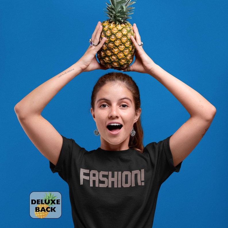 May include: A black short-sleeved t-shirt featuring the word FASHION! in a metallic, studded font across the chest. The garment is displayed against a solid blue background, with a small square logo in the corner reading DELUXE BACK.