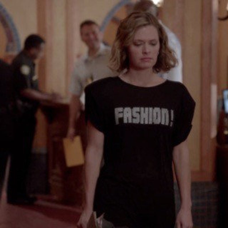 May include: A black short-sleeved t-shirt featuring the word FASHION! printed in large, bold, light-colored capital letters across the chest. The garment has a crew neckline and a relaxed fit with slightly dropped shoulder seams.