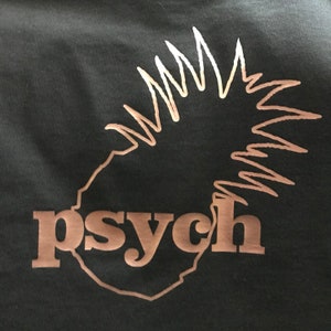 May include: A black t-shirt featuring a metallic copper-colored graphic of a pineapple silhouette. The word "psych" is printed in lowercase, bold, copper-colored letters across the lower portion of the pineapple design on the front of the shirt.